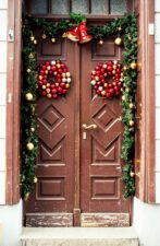 Puerta doble marrón enmarcada por guirnaldas luminosas con dos coronas de bolas de Navidad rojas y doradas, complementadas con grandes campanas rojas, irradiando un espíritu festivo y acogedor.